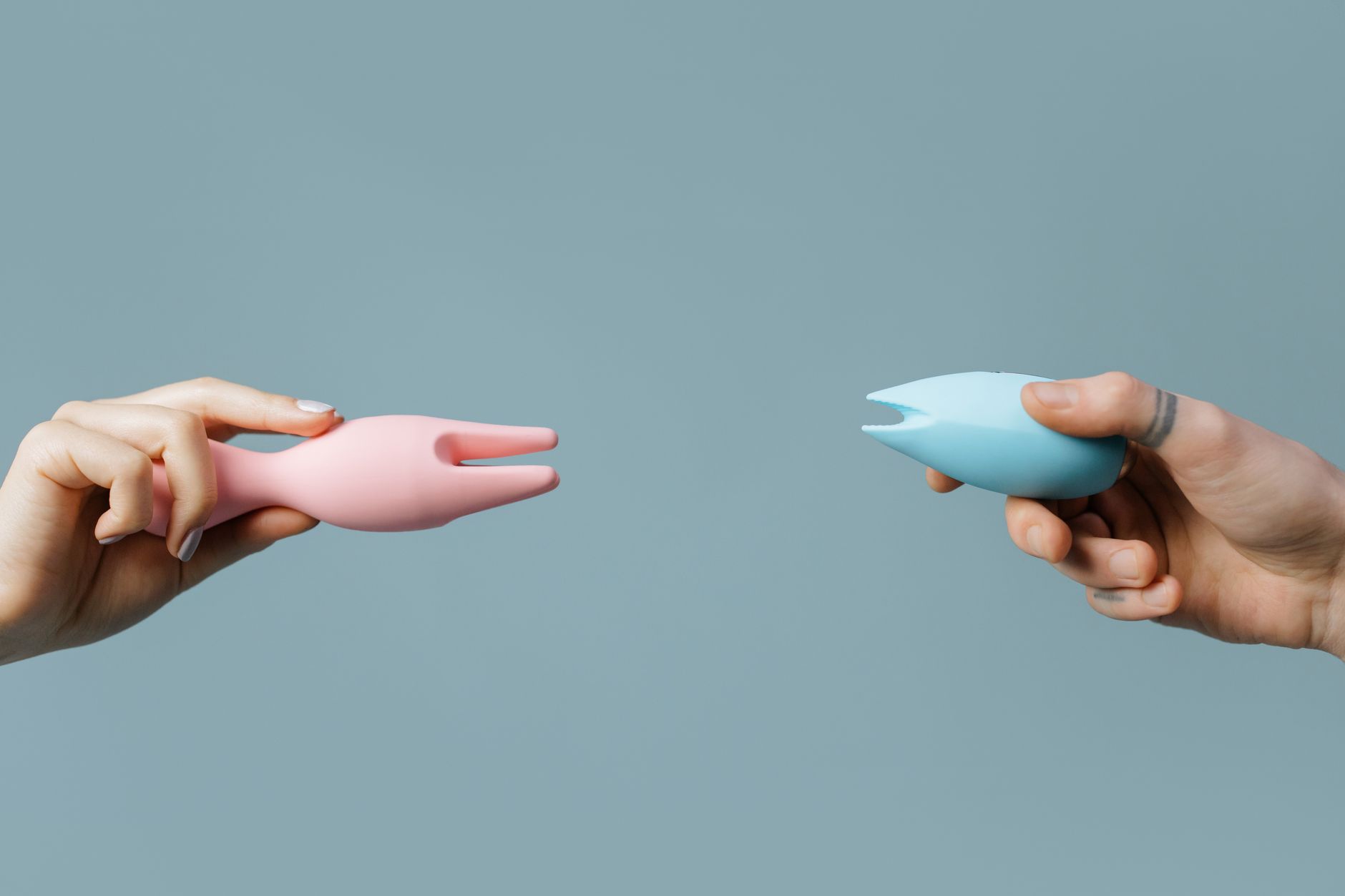Two hands holding clitoral vibrators against a pastel background, representing different options for managing sensitivity