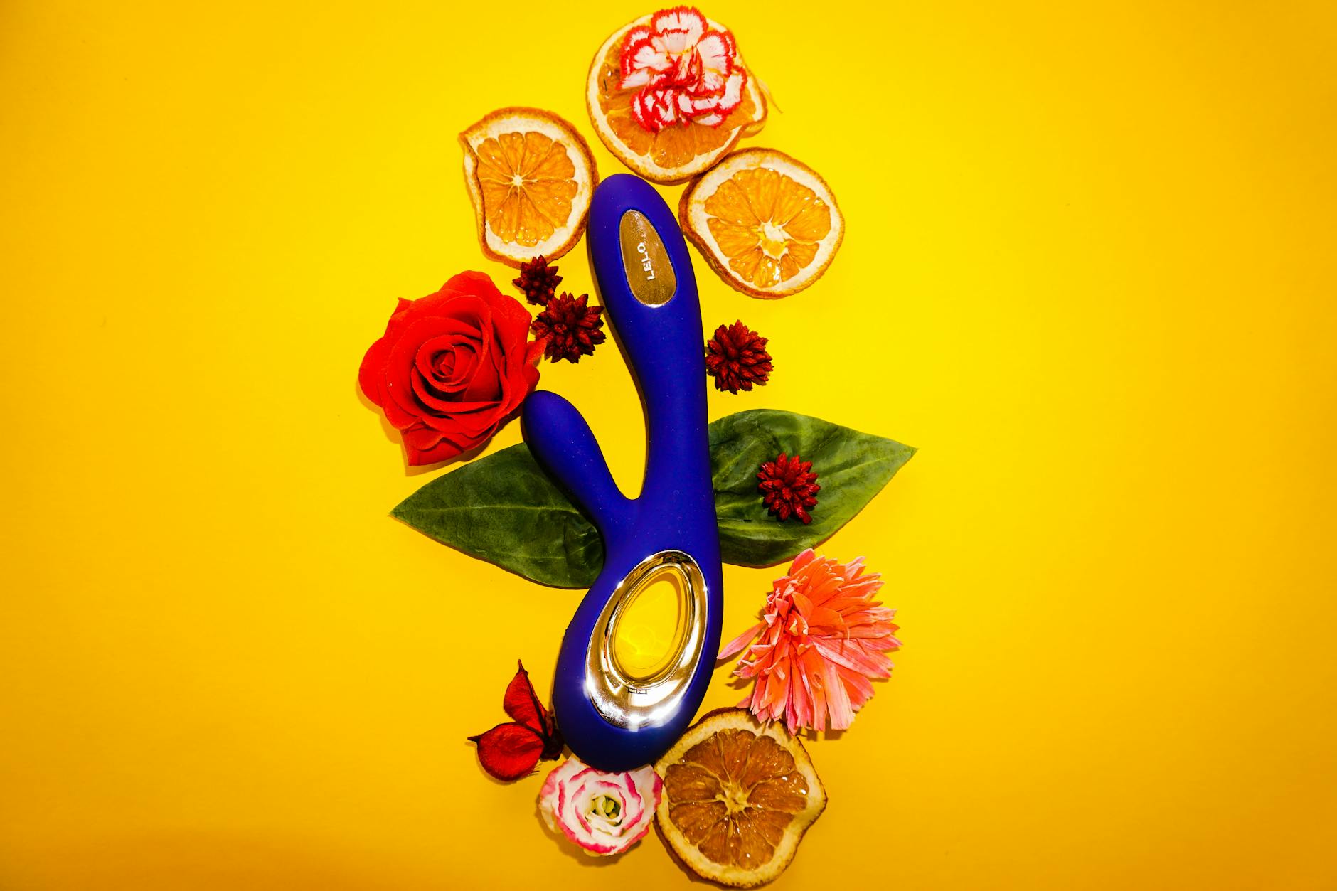 A blue vibrator surrounded by flowers and citrus on a bright yellow background symbolizing joy and renewal